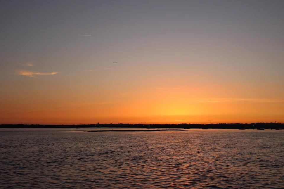 A serene sunset over a calm body of water with an orange and purple sky, and distant silhouettes of land and buildings