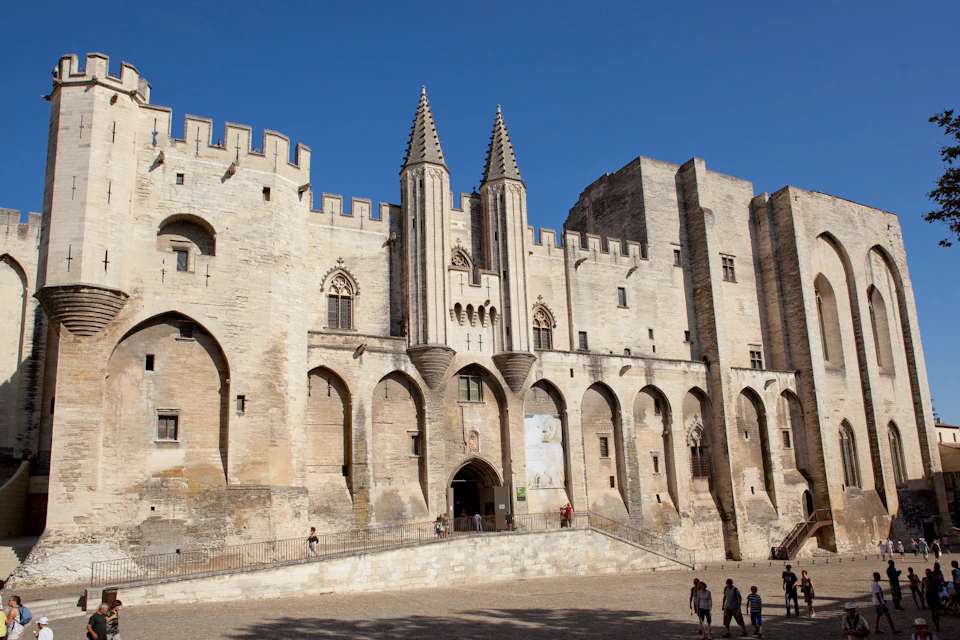 Historic stone building with tall towers, arched entrances, and a battlement top. A few people walk in the foreground. Clear sky.