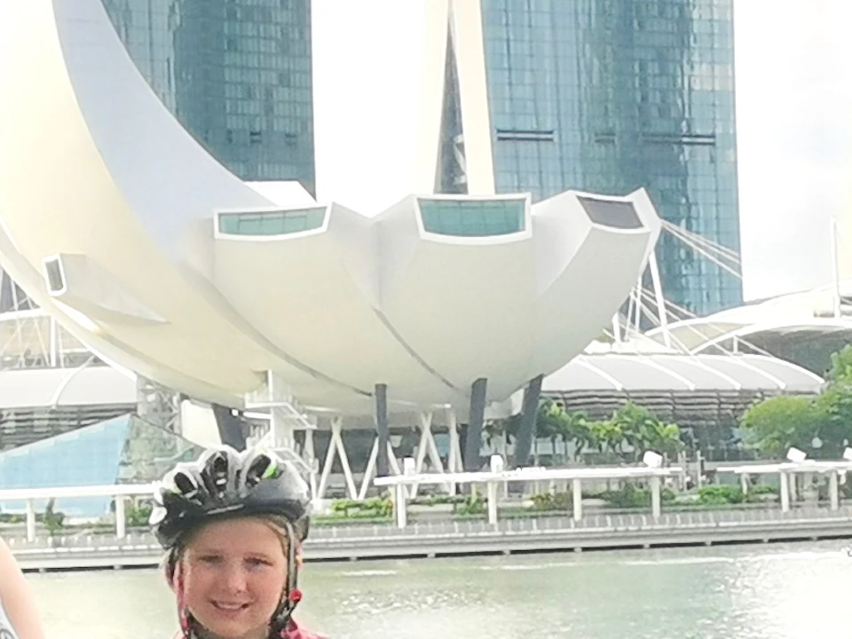 Six people with bicycles and helmets pose by the waterfront with Marina Bay Sands and a stadium in the background.