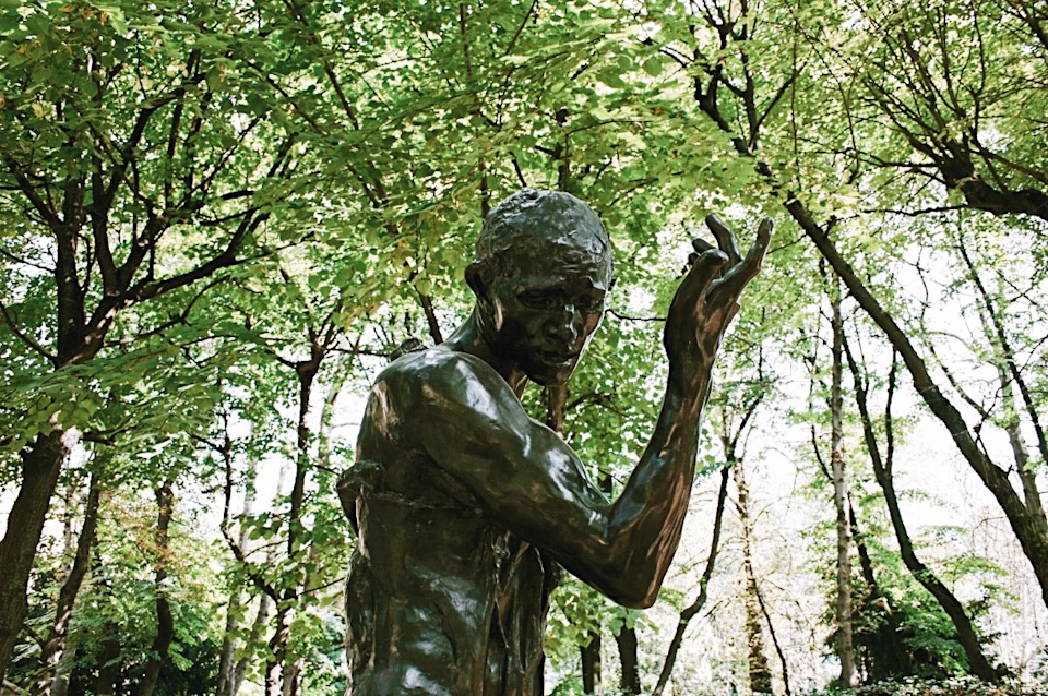 Bronze statue of a standing figure with raised arms, set against a background of lush green trees.