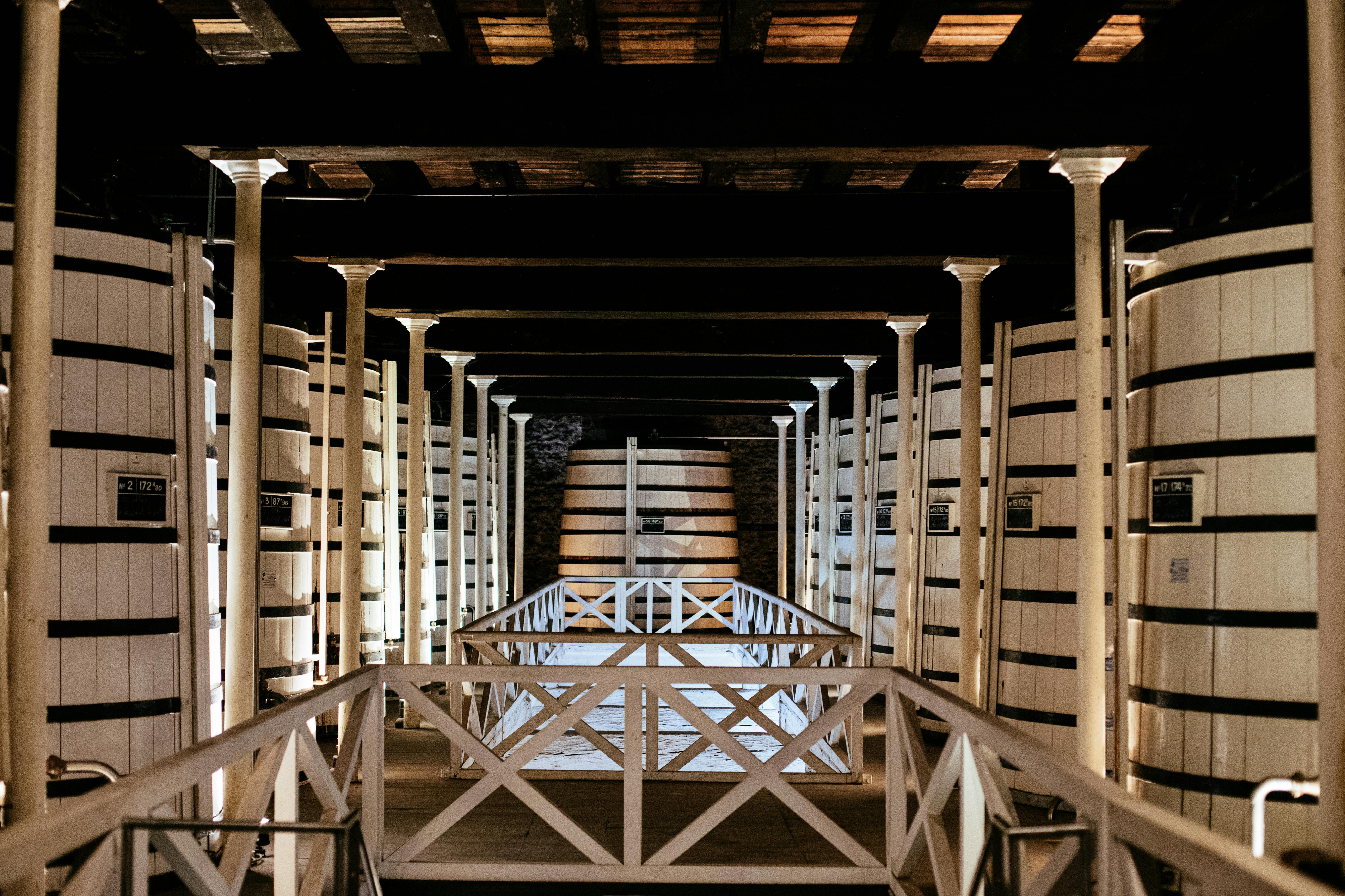 A walkway with white railings leads through a dim, wooden-beamed room with large, white barrels lined up on both sides.