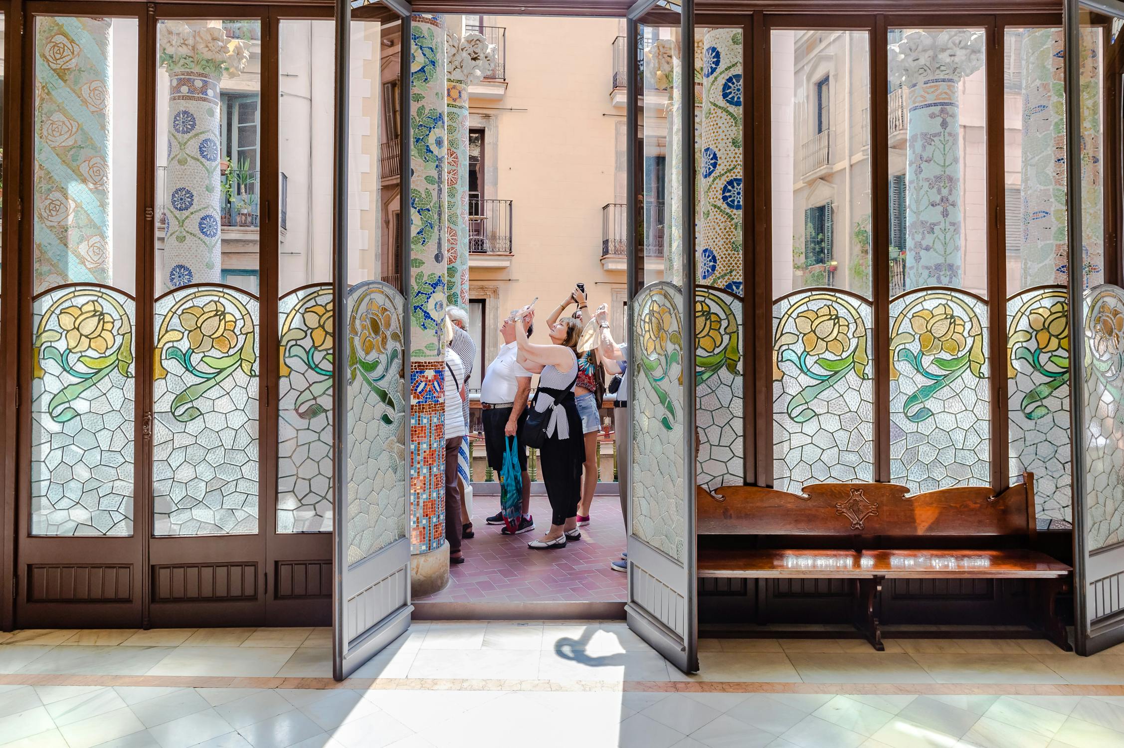 Eine Gruppe von Menschen fotografiert durch offene Mosaiktüren mit bunten Fliesen und Glasmalereien, die zu einem Korridor im Freien führen.