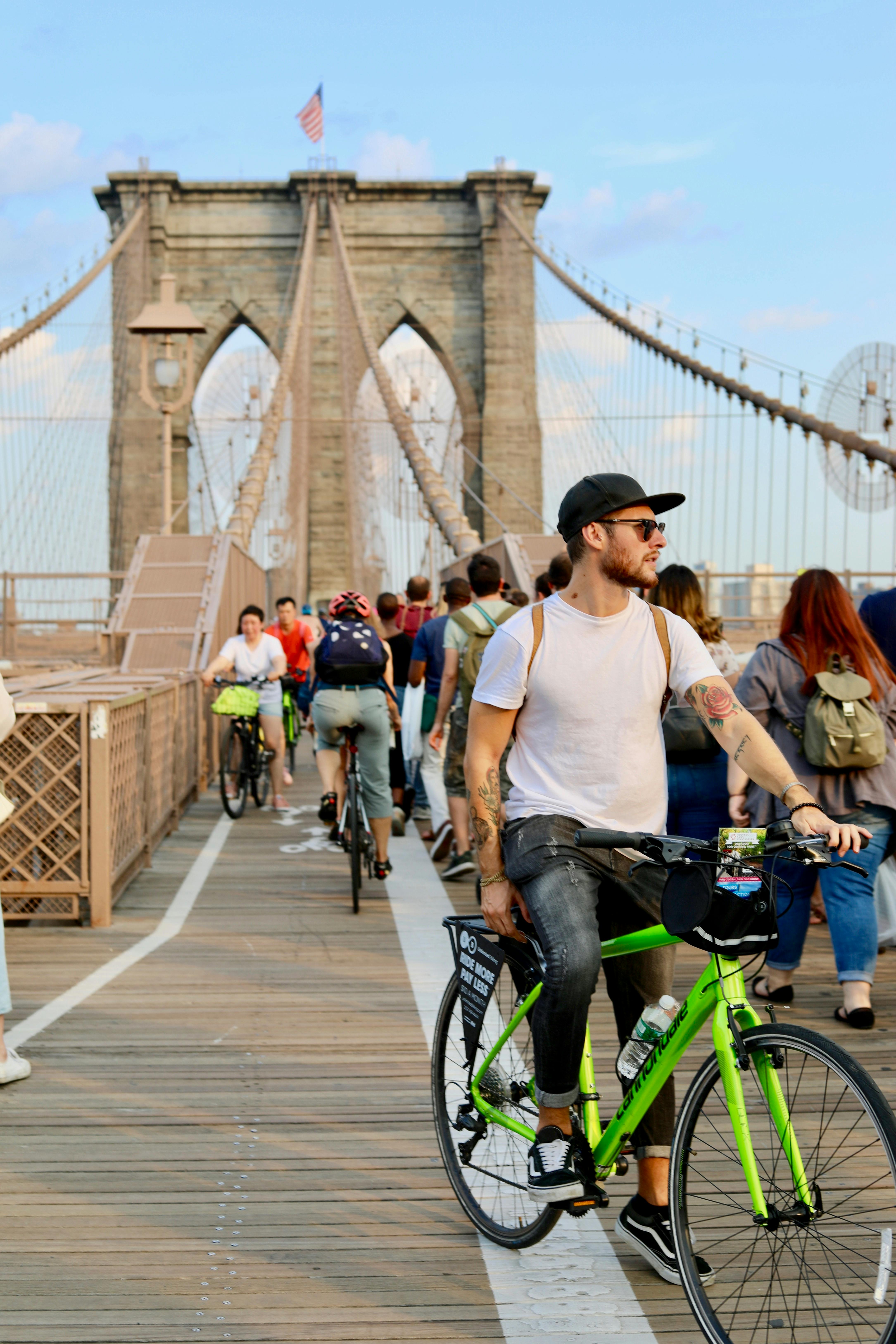 Persone in bicicletta e a piedi sul ponte di Brooklyn sotto un cielo limpido, con le torri di pietra del ponte sullo sfondo.