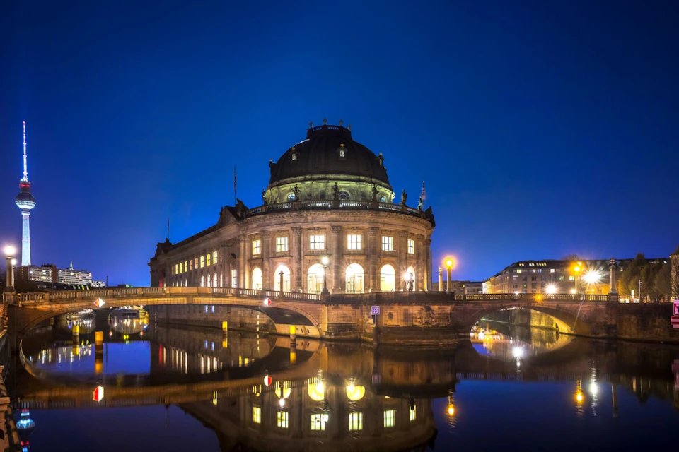 Illuminated domed building with arched windows reflecting on a river at night under a clear, dark sky.