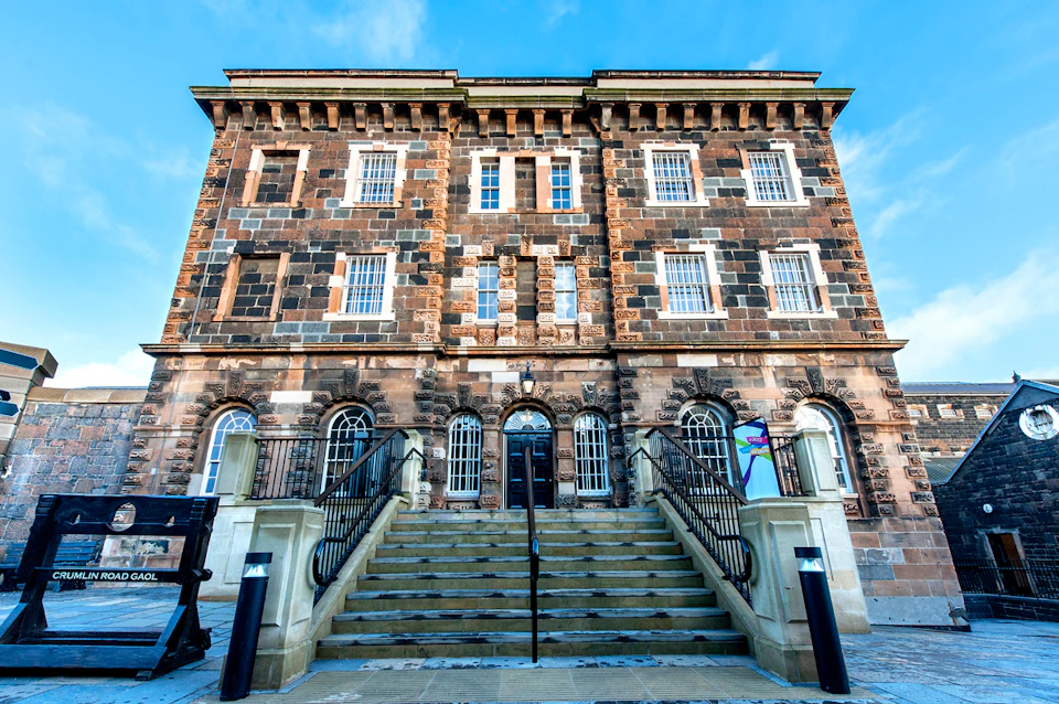 A three-story, historical stone building with stairs leading to the entrance, featuring arched windows and a bright blue sky background.