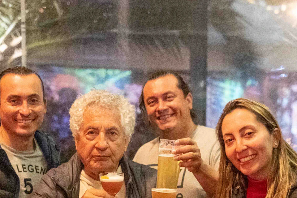 A group of five people sitting at a table, smiling and holding glasses of beer in a warmly lit indoor setting.