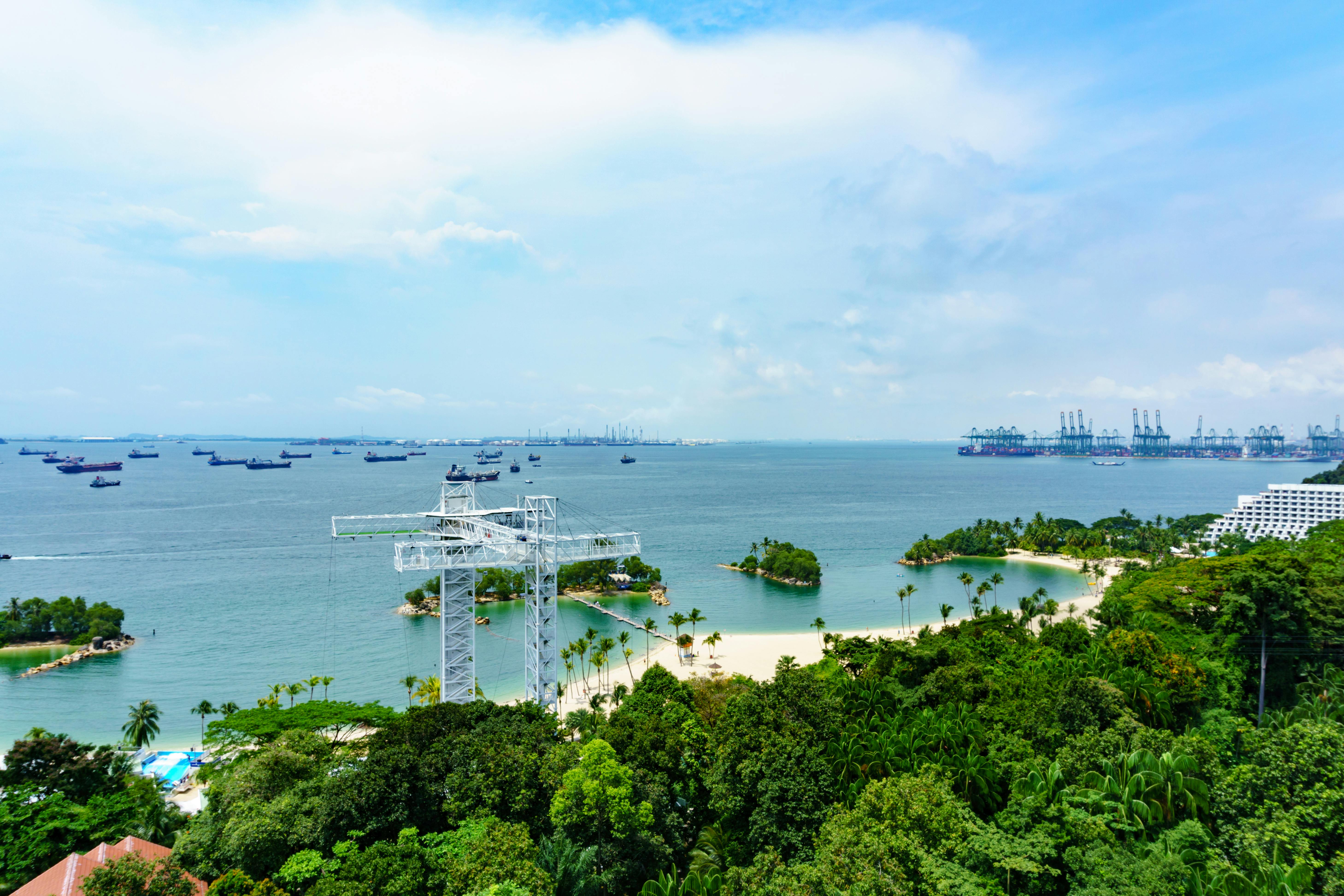 Aerial view of a coastal area with dense greenery, a white metal structure, sandy beach, small islands, and numerous ships on the sea.