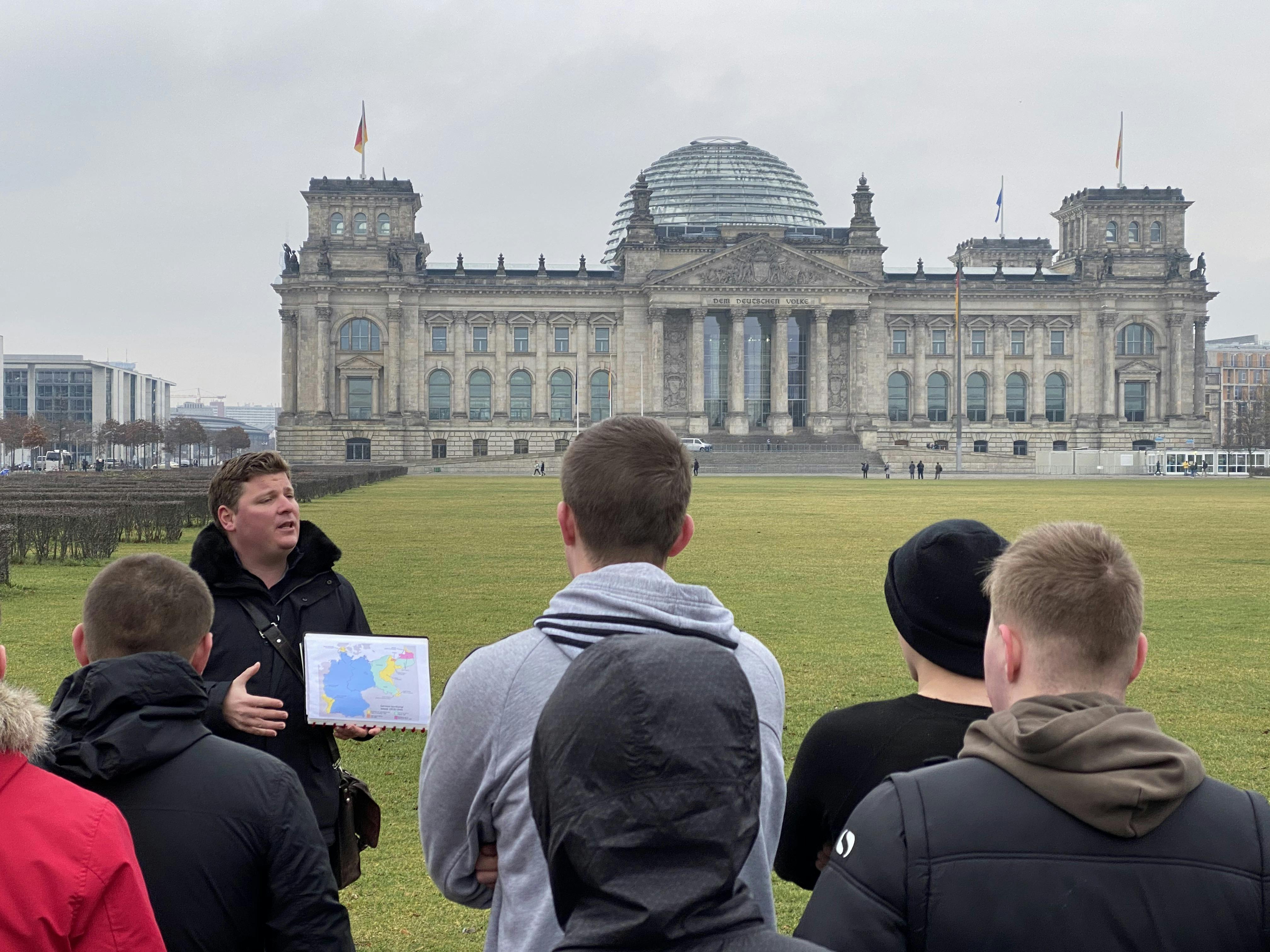흐린 날, 독일 베를린의 국회의사당 건물 밖에서 한 무리의 사람들에게 지도를 보여주는 사람.