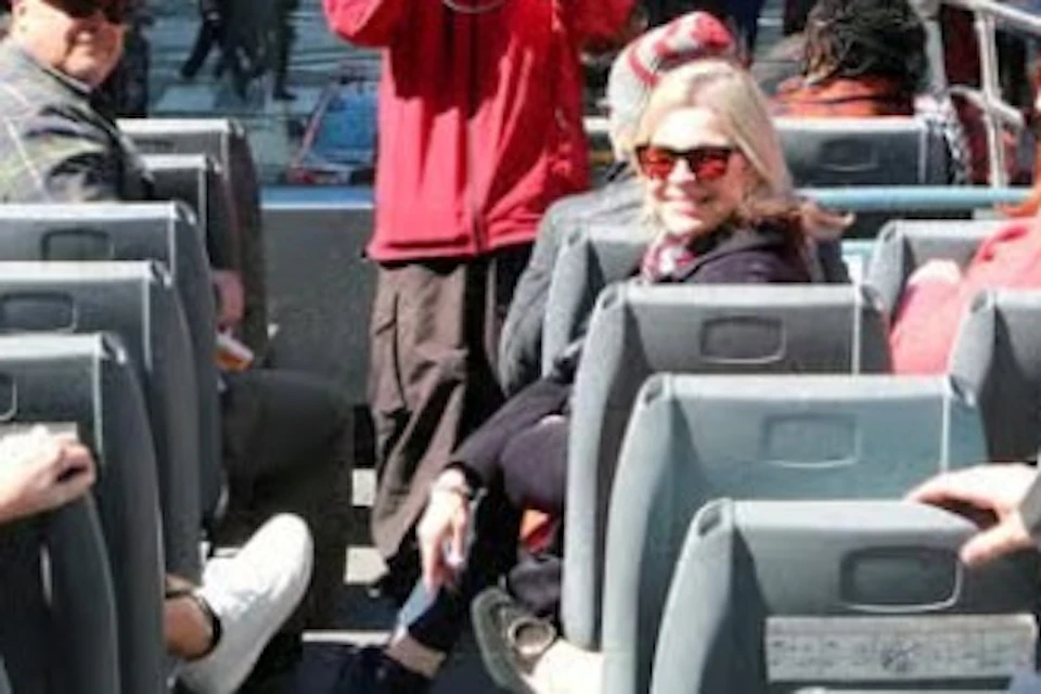 A group of people on an open-top tour bus in a city, with urban buildings and signs visible in the background.