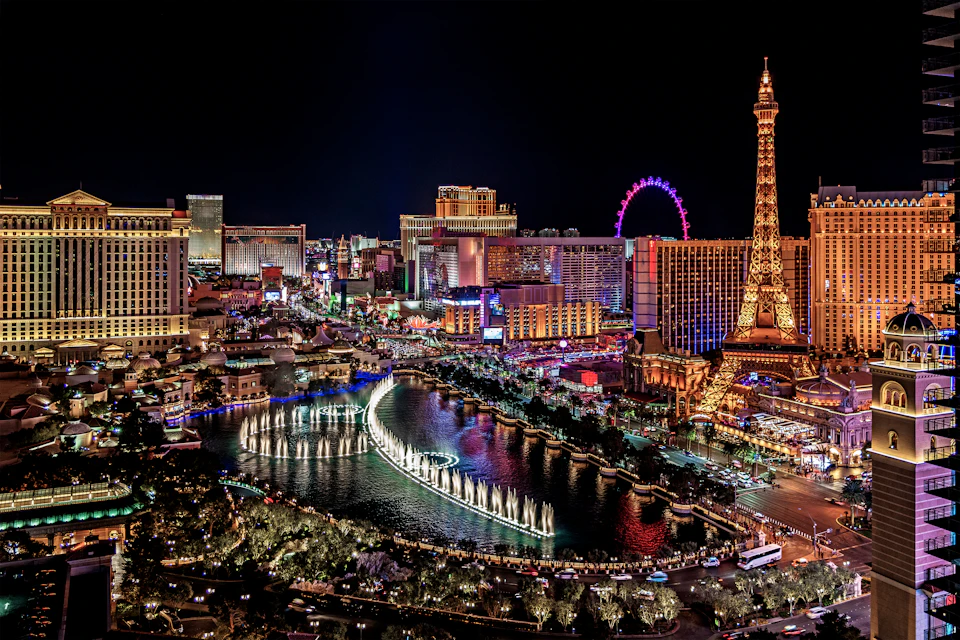 Nighttime aerial view of Las Vegas Strip with illuminated buildings, Eiffel Tower replica, Ferris wheel, and fountain display.