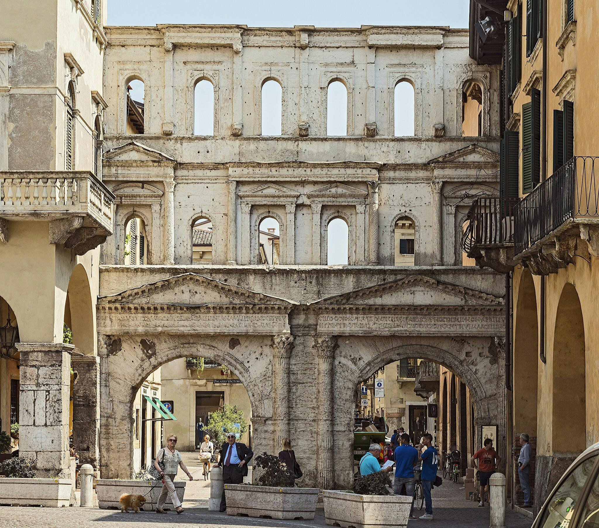 Antico arco in pietra con finestre multiple circondato da negozi ed edifici. Persone che camminano e conversano nella strada sottostante.