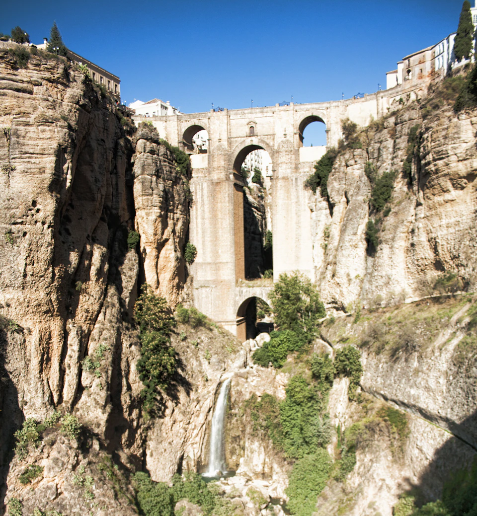 Stone bridge with three arched spans over a deep gorge with a waterfall; buildings line the top of the canyon. Clear blue sky.