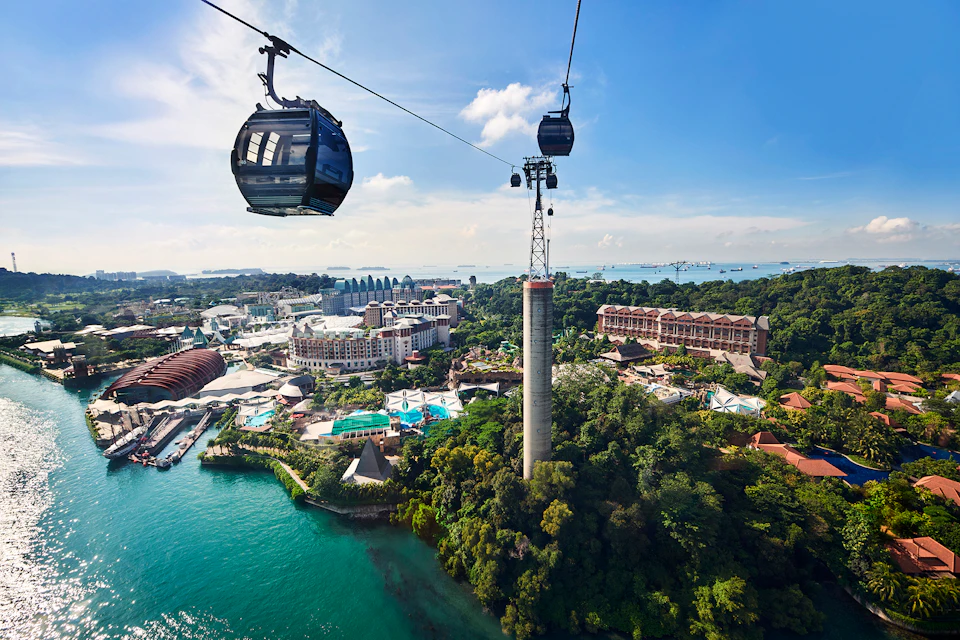 Two cable cars travel over a lush island with resorts, pools, and other buildings. The sea and additional islands are in the background.
