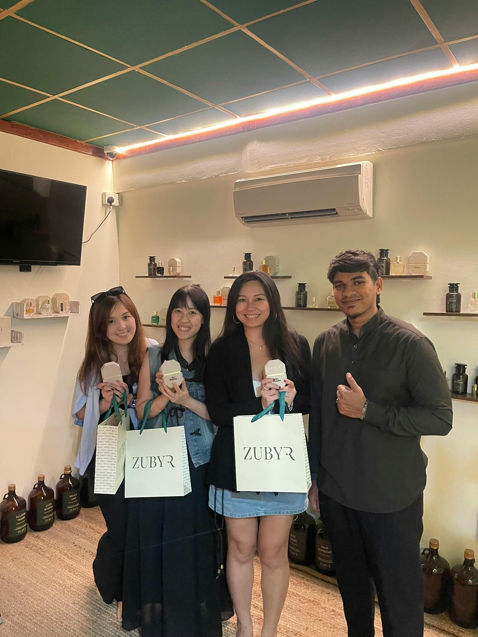 Four people stand indoors, three women holding Zubyr bags and products, and one man gesturing positively. Shelves with items are behind them.