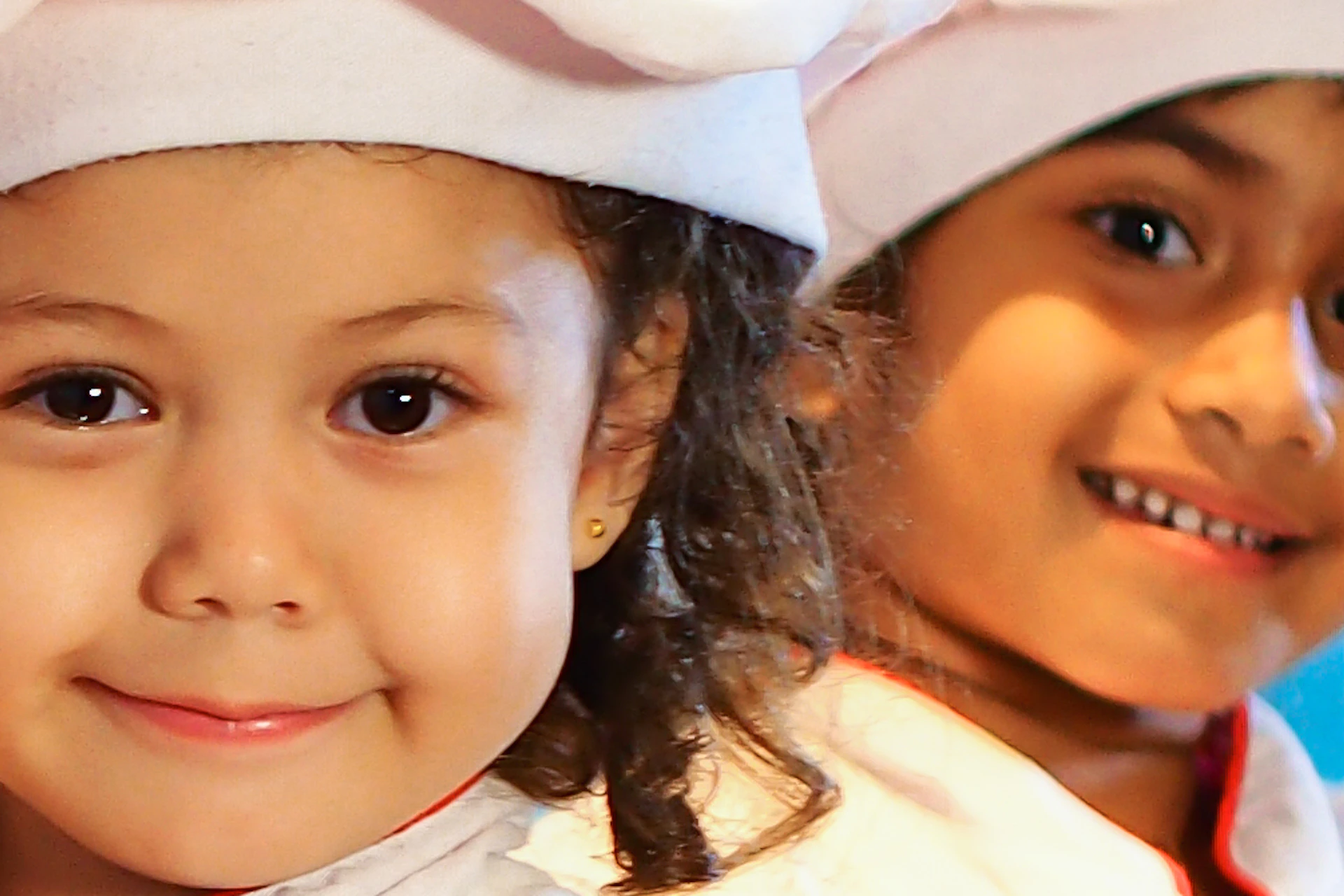 Two children wearing chef outfits hold toy pans in a play kitchen with colorful tile backsplash and motivational posters.