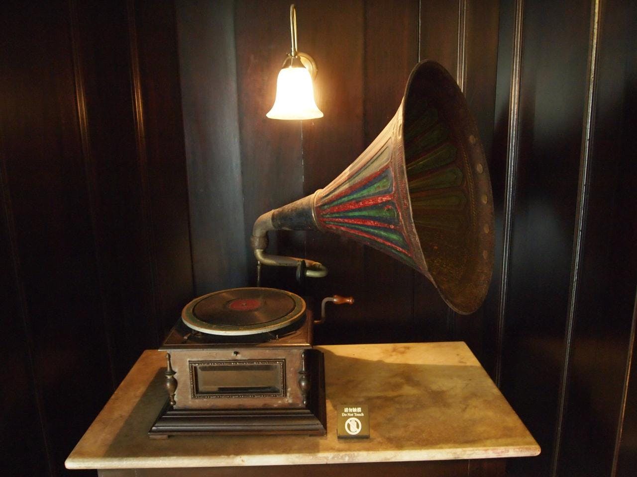 Starožitný gramofon s velkým barevným rohem sedí na stole pod malou osvětlenou nástěnnou lampičkou.
