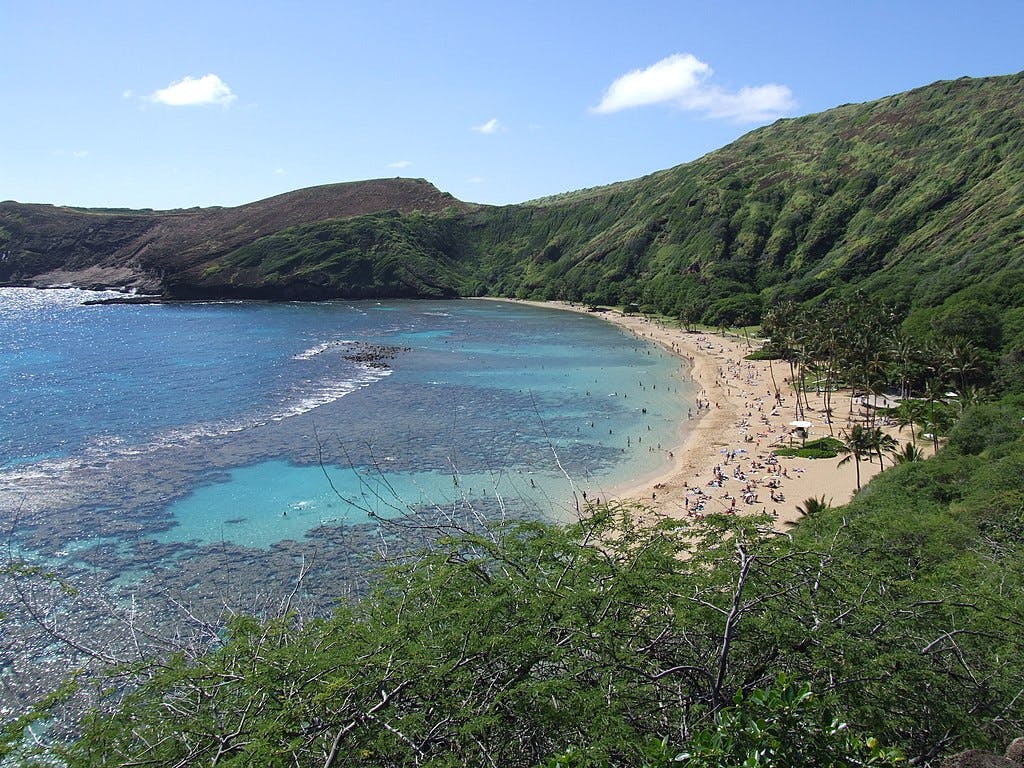 Sandy beach with people, turquoise water, coral reefs, lush green hills, and clear blue sky with a few clouds.
