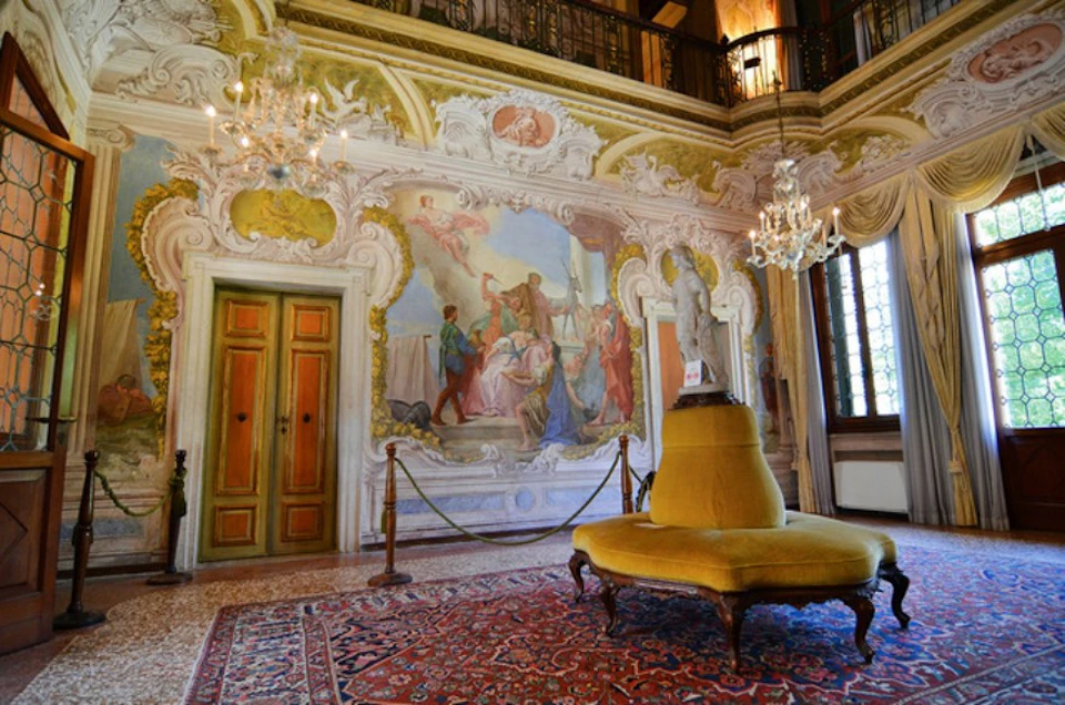 Ornate room with frescoed walls, chandeliers, a yellow upholstered bench, and a patterned rug. A statue stands in the center.