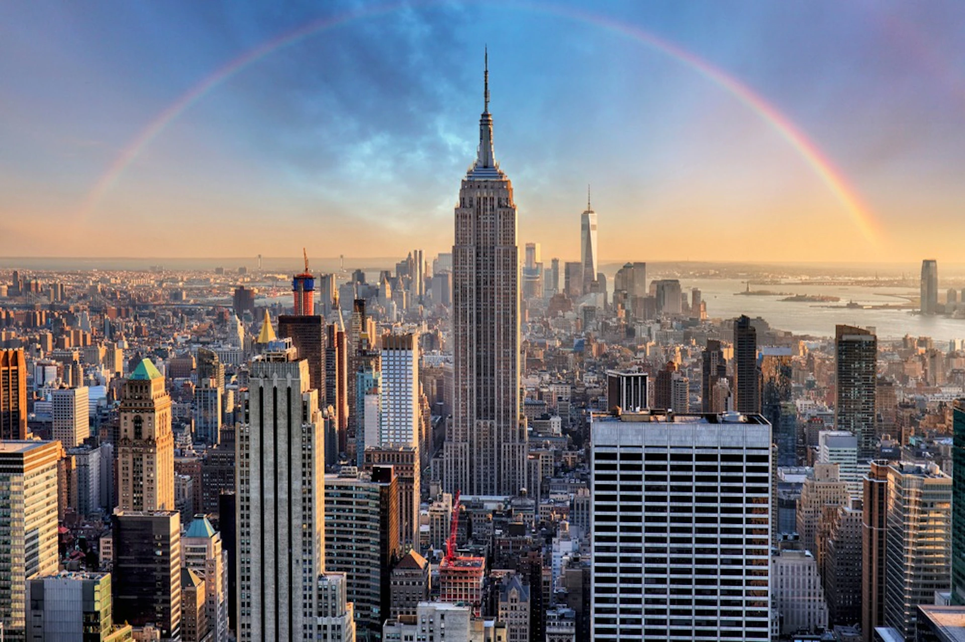 Cityscape featuring the Empire State Building with a rainbow arching over the skyline at sunset.
