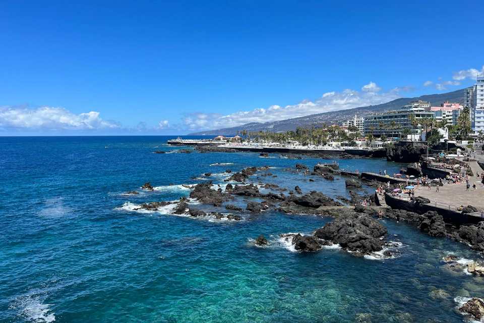 Rocky coastline with clear blue ocean water, palm trees, and buildings in the background under a bright blue sky with scattered clouds.