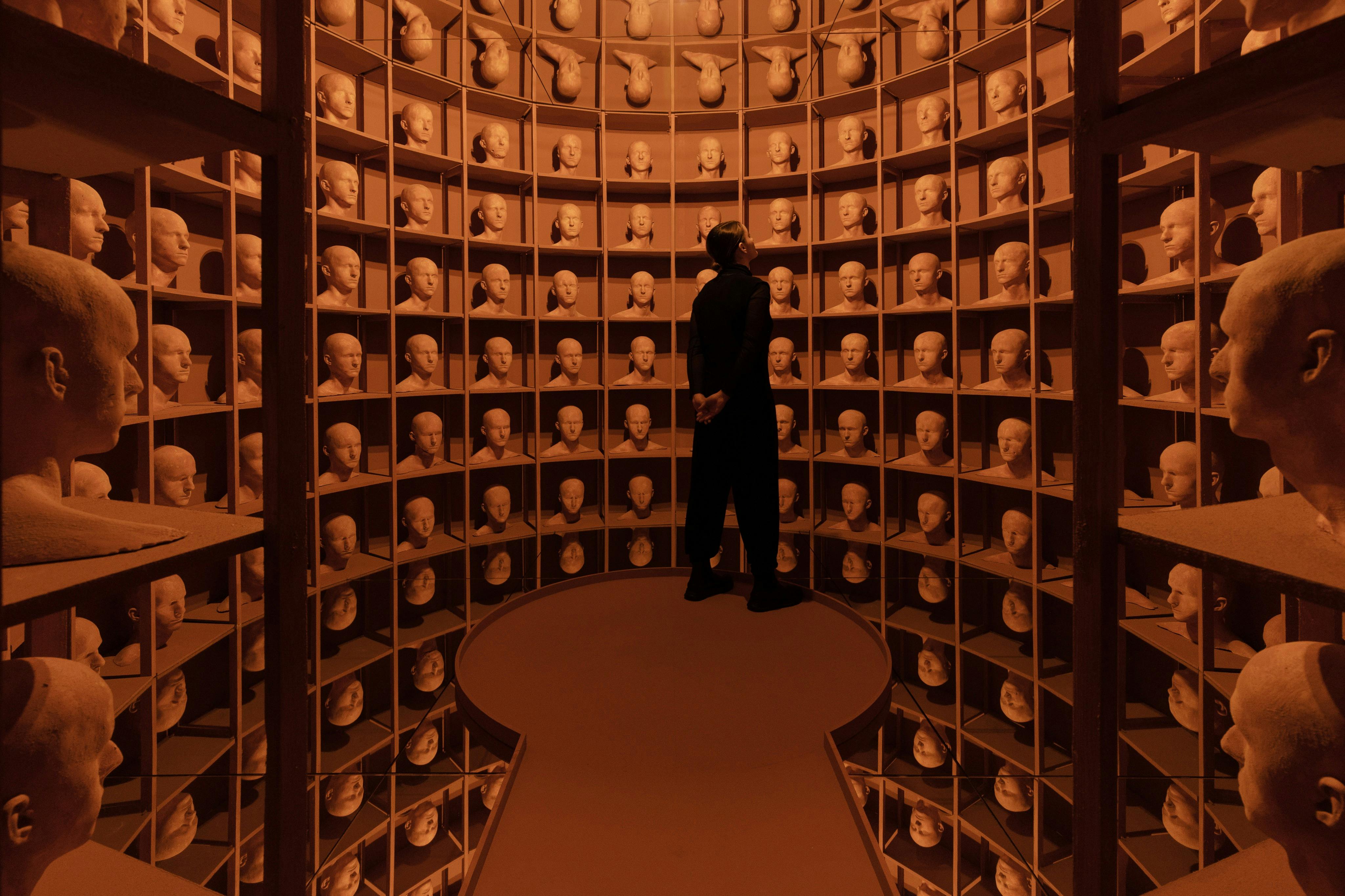 동일한 머리 조각이 늘어선 선반과 주황색 조명이 켜진 원형의 방에 서 있는 사람.
