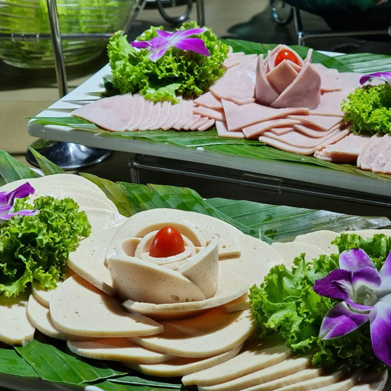 Dva talíře nakrájeného delikatesního masa s ozdobami hlávkového salátu, fialovými květy a cherry rajčaty na banánových listech.