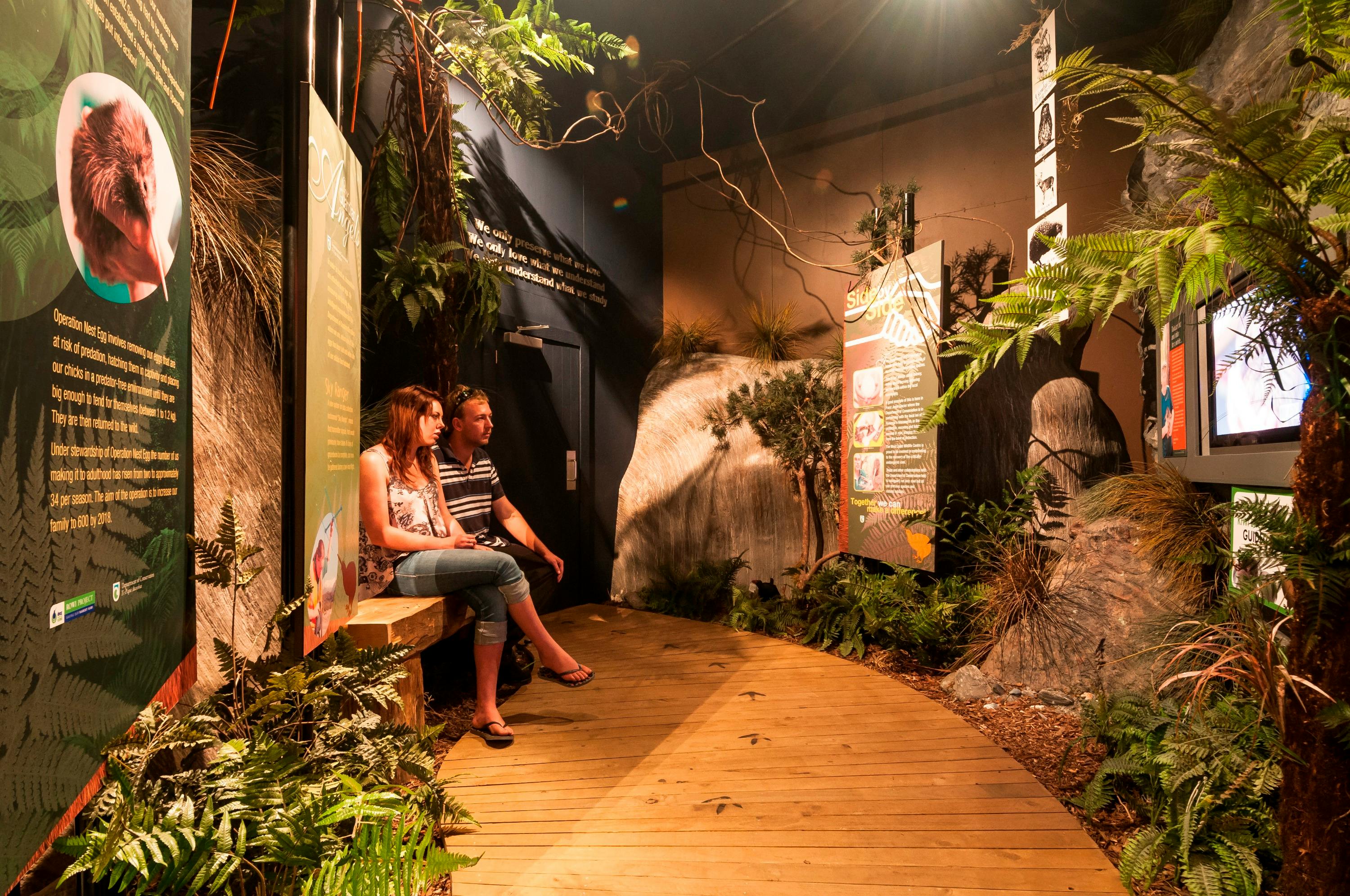 Two people sitting on a bench in an indoor exhibit with tropical plants, informational panels, and a wooden walkway.
