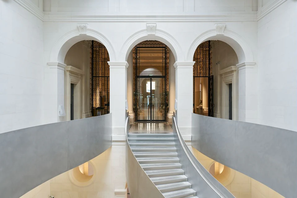 A grand indoor staircase with sleek railings, leading to a hall with arched doorways and ornate glass and metal doors.