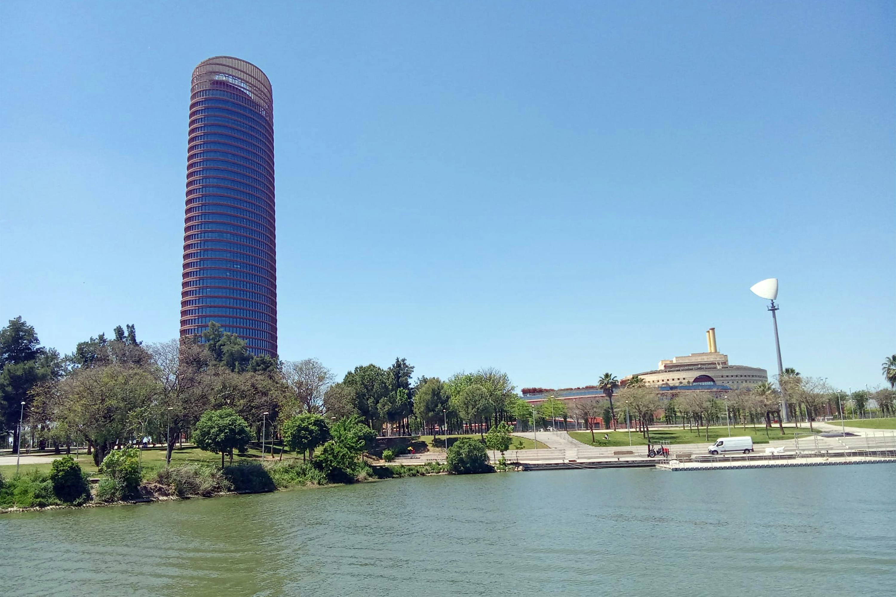 Un alt edifici cilíndric de color vermell i vidre s&#39;alça prop d&#39;un parc amb arbres i un riu, amb un altre edifici únic al fons.