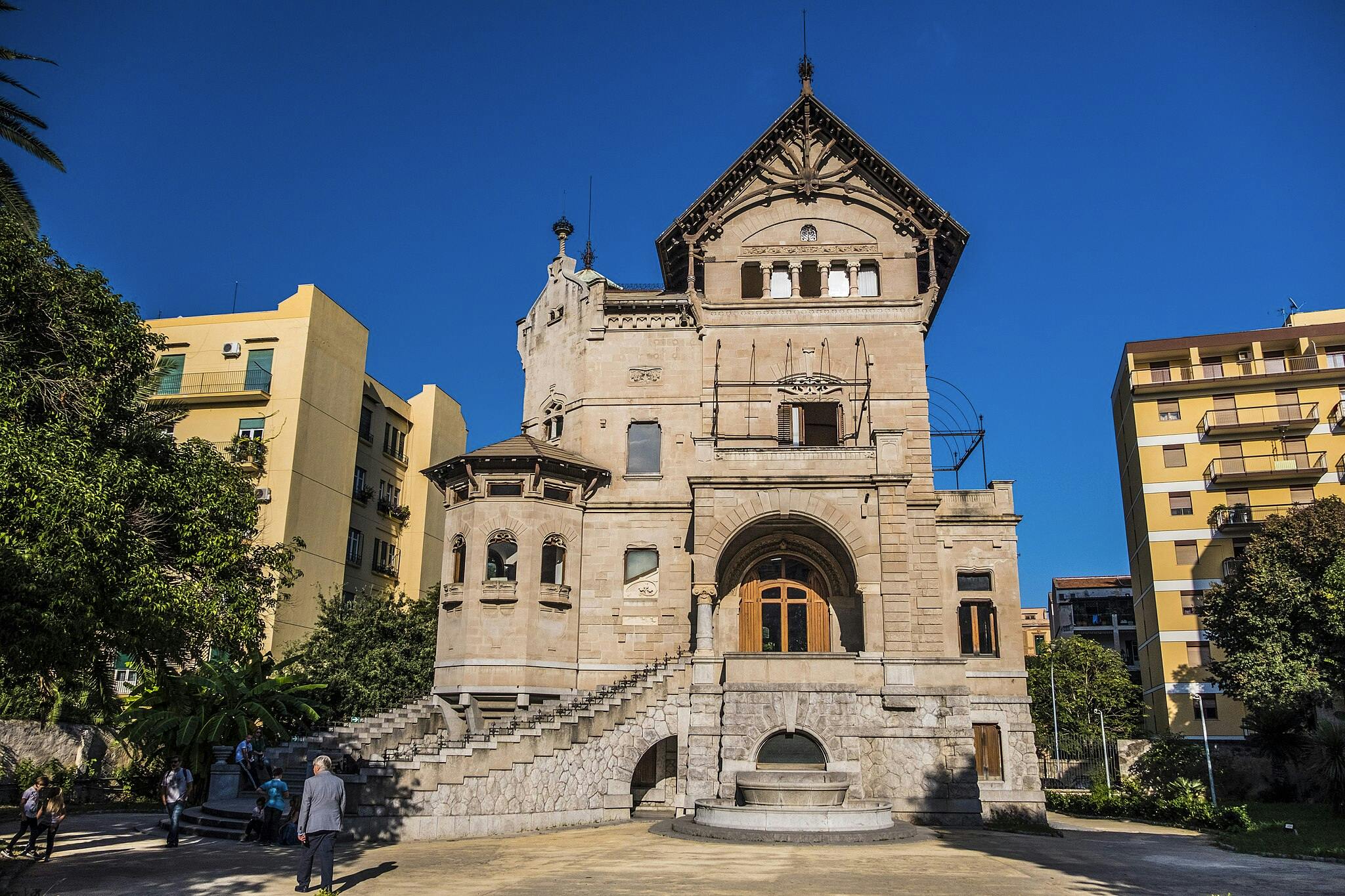 Ornate, historic building with arched entrance and curved steps. Modern yellow buildings and trees are in the background.