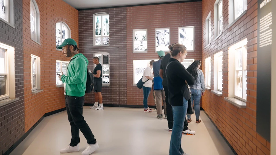 A group of people observing displays in a brick-walled exhibit room with illuminated panels and photos on the walls.