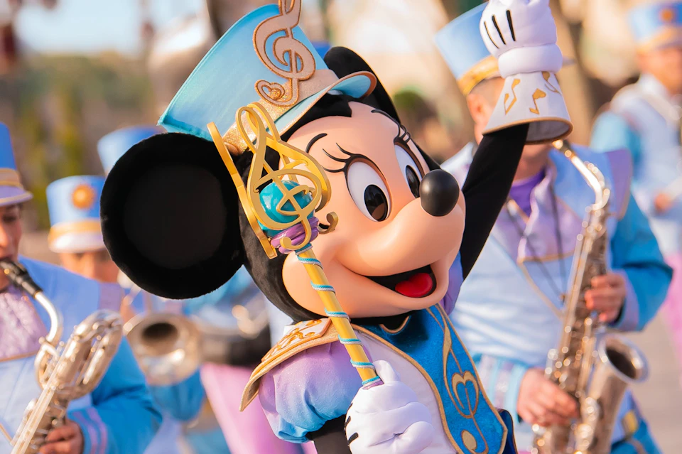 A costumed character wearing a musical-themed outfit and holding a staff, taking part in a parade surrounded by performers.