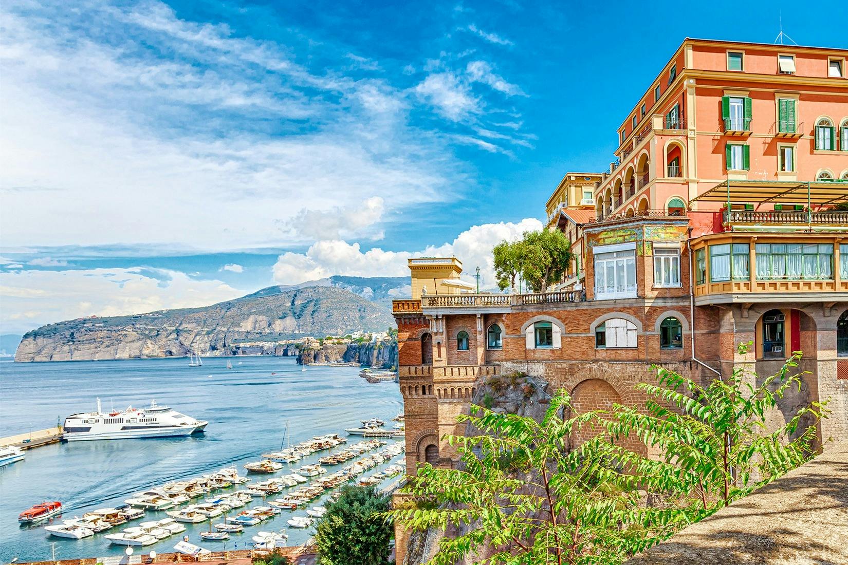 崖の上に大きな建物があり、マリーナと山の背景に停泊しているボートを見下ろすウォーターフロントのシーン。