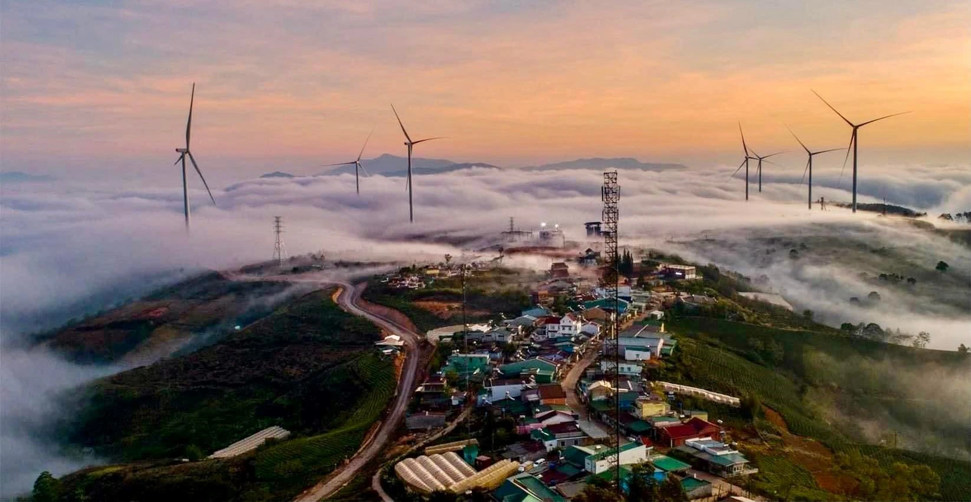 A scenic view of a village with colorful houses, wind turbines, and fog covering distant hills during a sunset.
