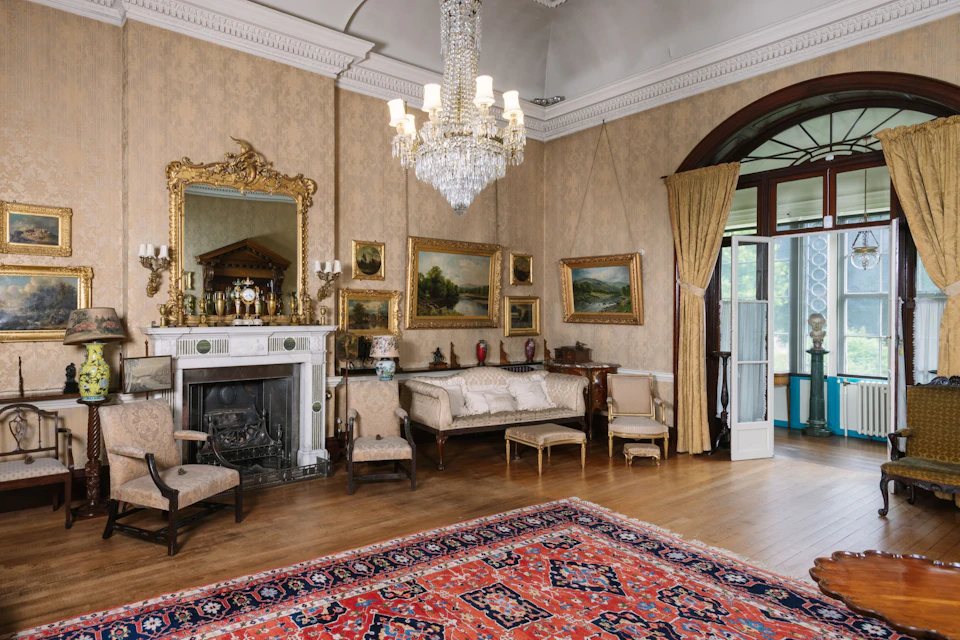 Elegant vintage living room with chandelier, ornate mirror above a fireplace, antique furniture, and framed art on the walls.