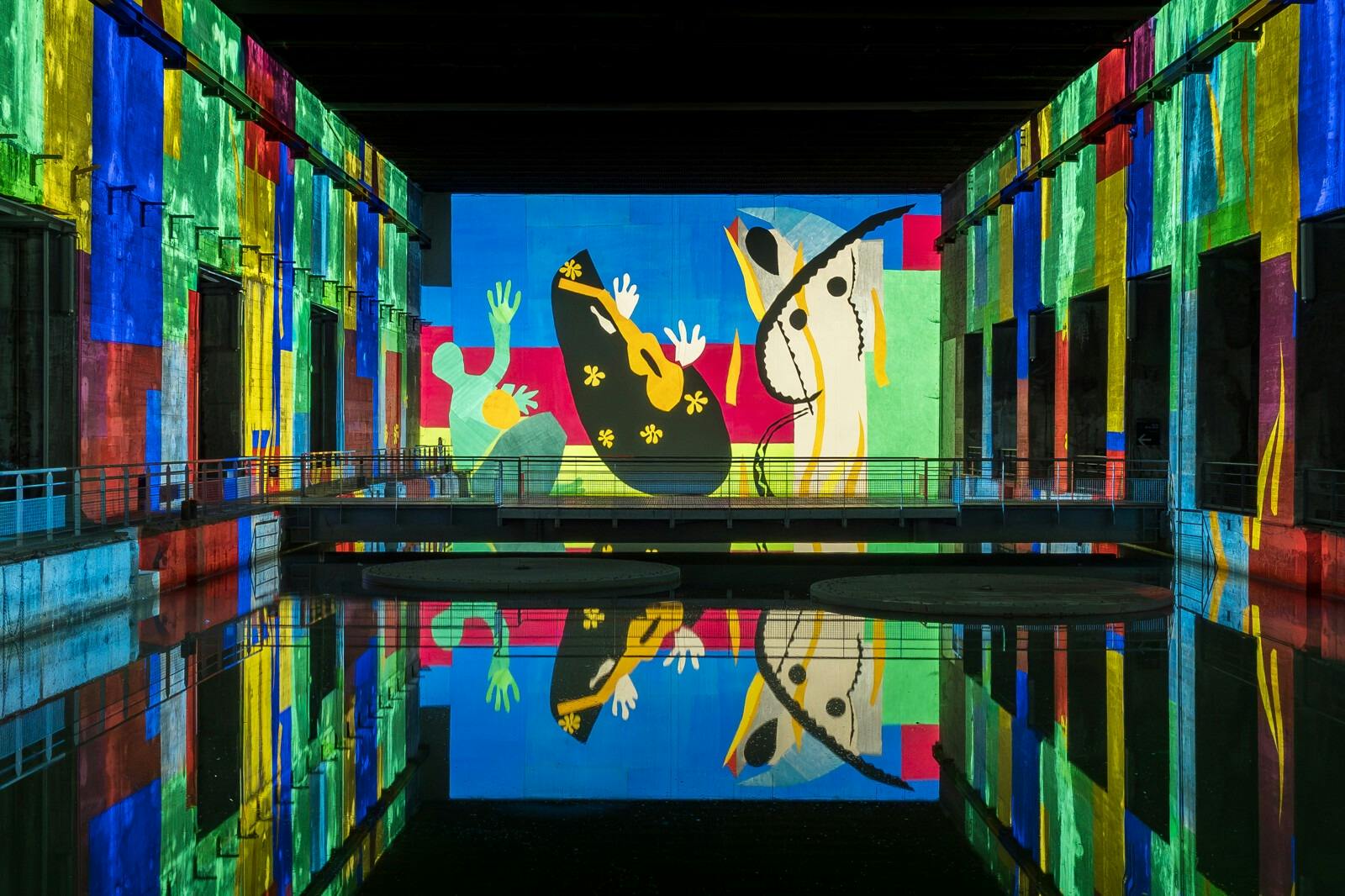 En una pared, sobre una superficie de agua reflectante, se ve un mural abstracto y lleno de color, rodeado de estructuras de colores.