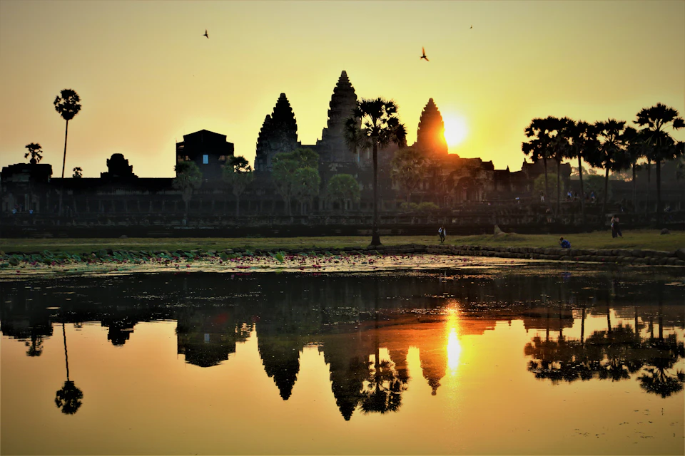 Silhouette of Angkor Wat temple at sunrise, with the sun partially visible behind the structure and a reflection in a lake.