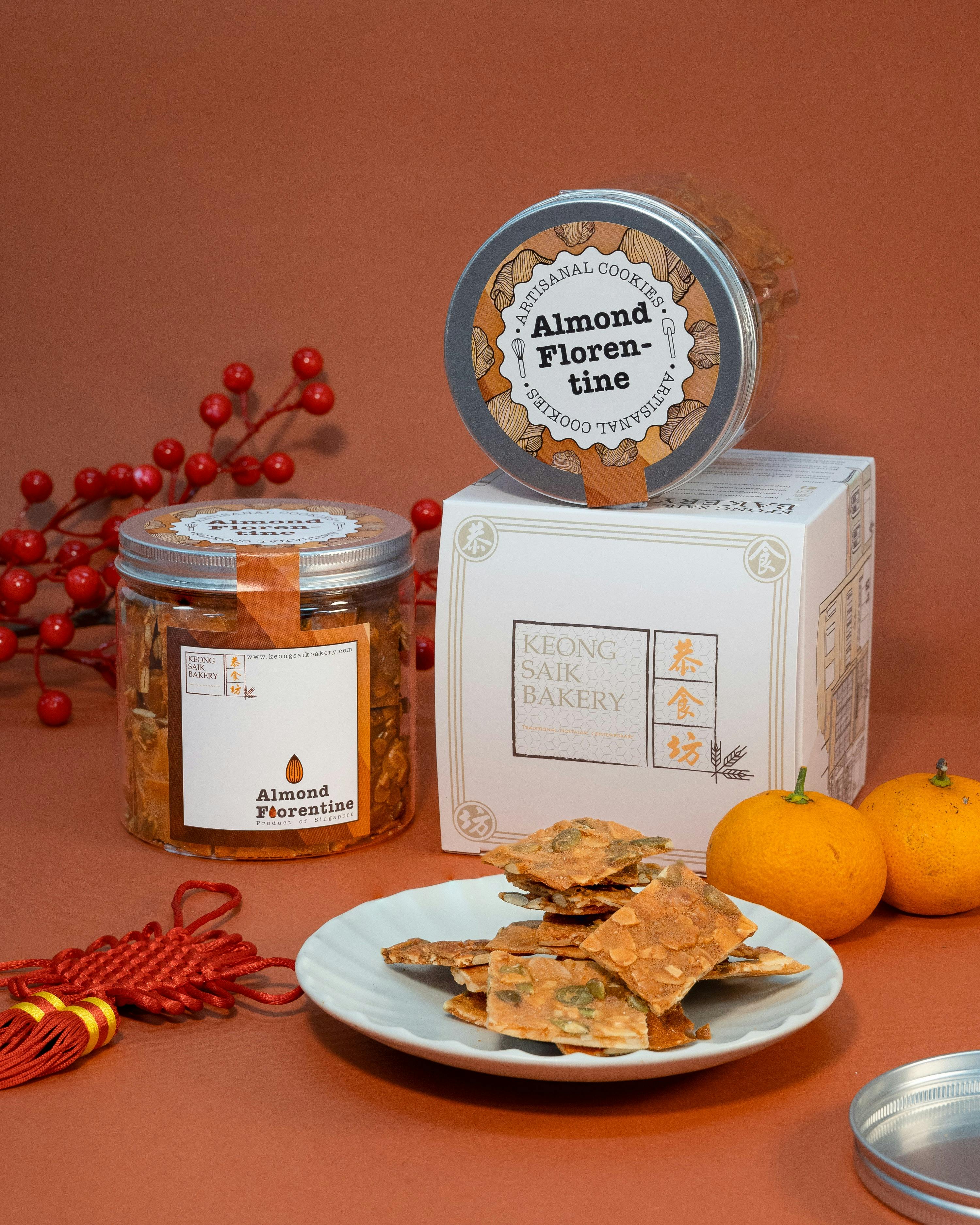 A display of almond florentine cookies in jars and a plate, with festive red decorations and oranges on a red background.