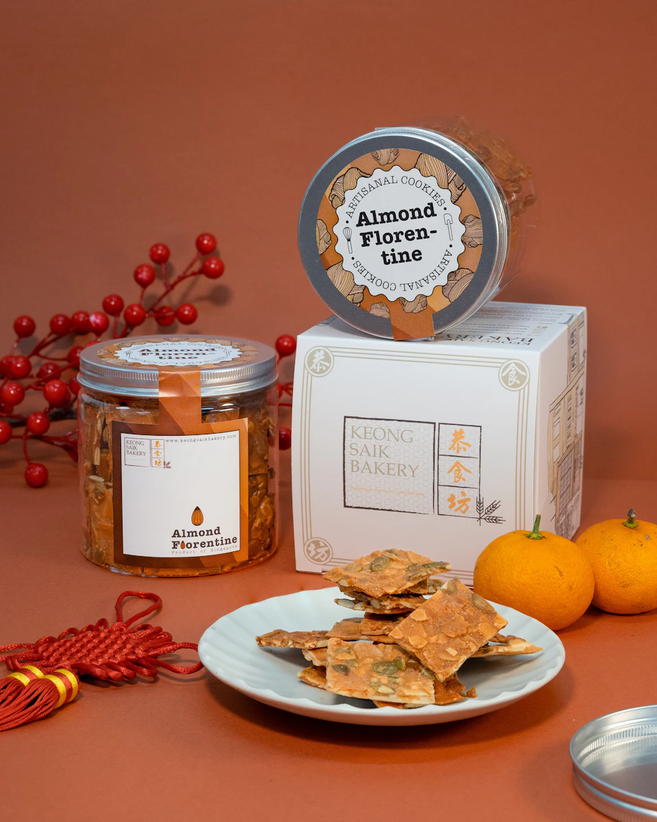 A display of almond florentine cookies in jars and a plate, with festive red decorations and oranges on a red background.