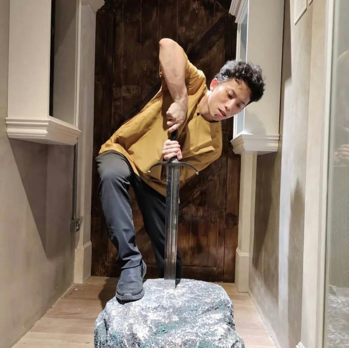 A person in a yellow shirt and dark pants pulls a sword embedded in a stone within a narrow hallway with a wooden door behind.