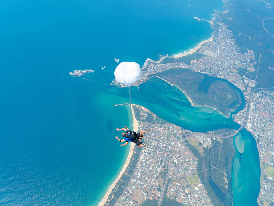 Two people are tandem skydiving, with a white parachute deployed above a coastal landscape featuring beaches, water, and buildings.
