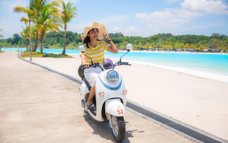 A person riding a white scooter by a beach, wearing a wide-brimmed hat and a yellow striped shirt, under a clear sky.
