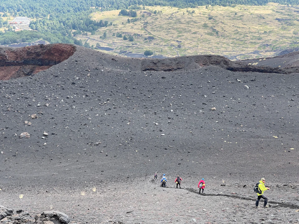 A group of people hiking up a rocky volcanic slope, surrounded by scattered stones, with a distant landscape in the background.
