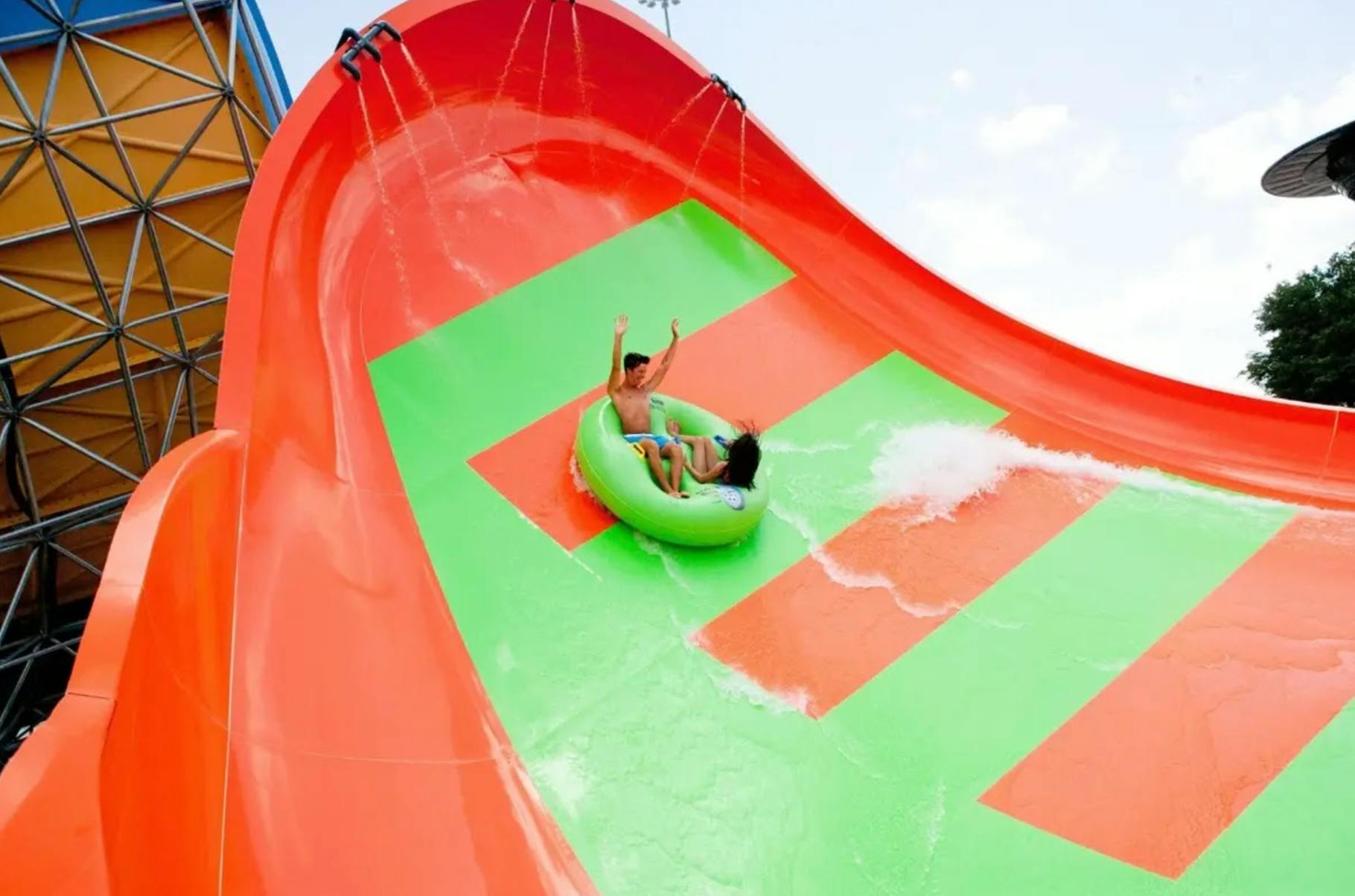 Dues persones pugen en un tub inflable verd per un tobogan d'aigua de colors taronja i verd brillants amb aigua que salpica.