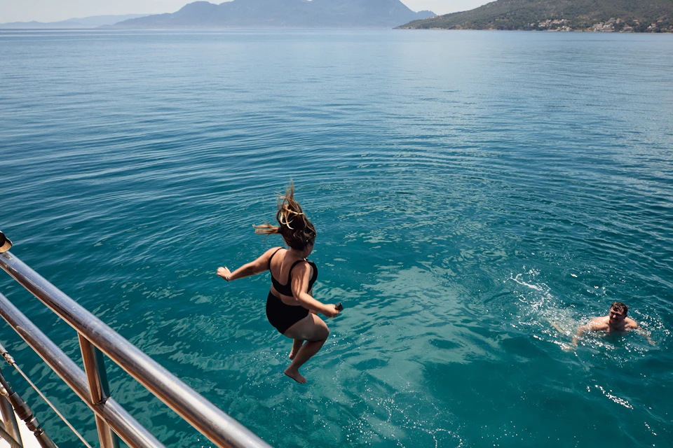 Person in a black swimsuit jumping from a boat into clear blue water, with islands in the background.