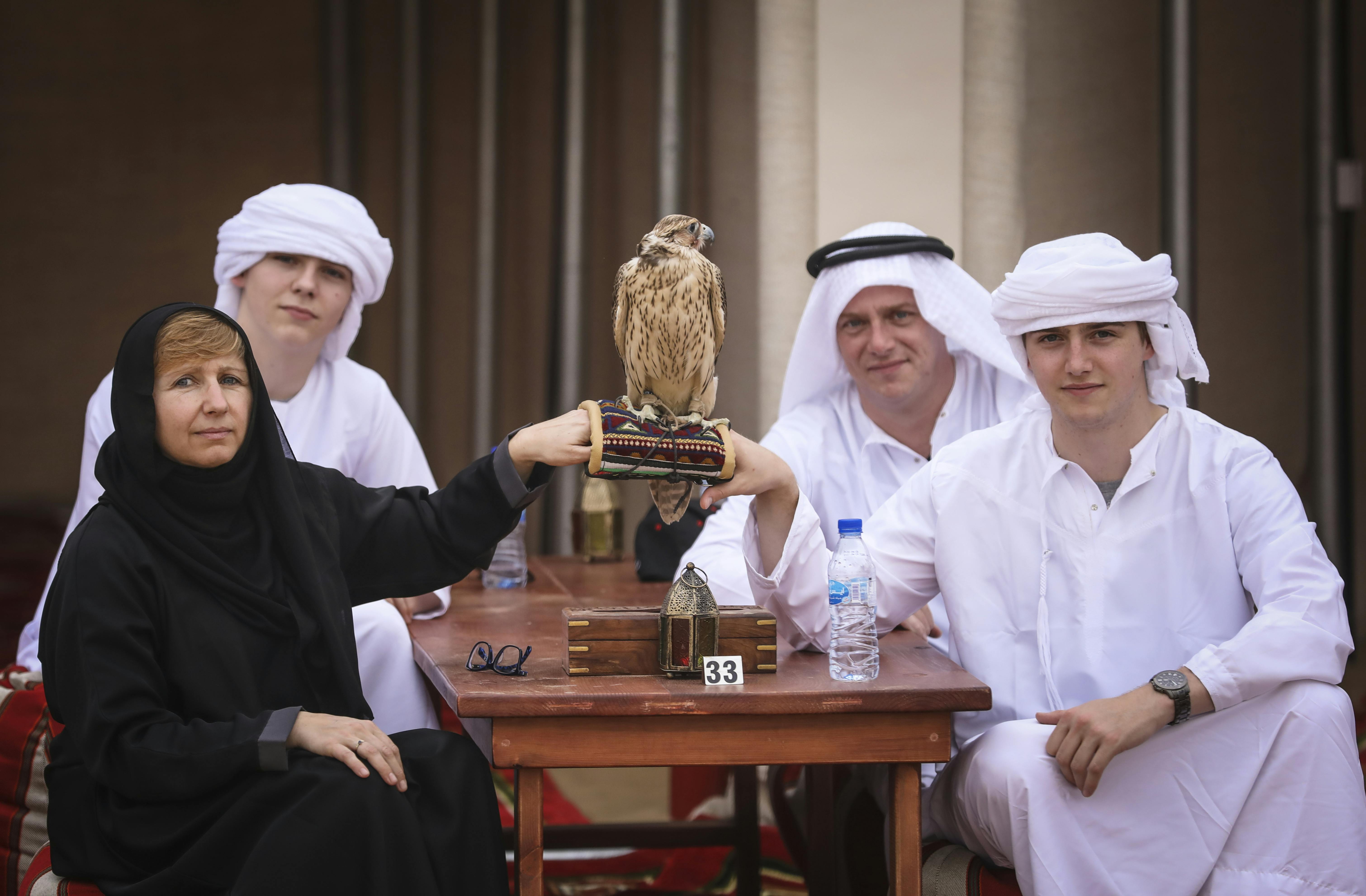 U stolu sedí tři lidé se sokolem posazeným na ruce ženy v rukavici. Na stole je láhev na vodu a sklenice.