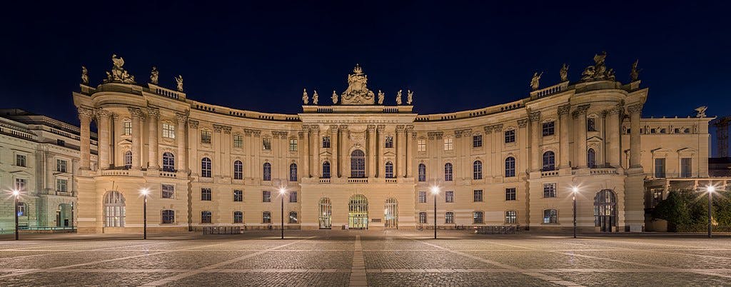 Illuminated historic building with ornate architecture and statues on the roof, seen at night with a wide, empty courtyard in front.