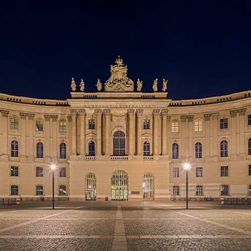 Upplyst historisk byggnad med utsmyckad arkitektur och statyer på taket, sett på natten med en bred, tom innergård framför.