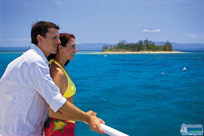A couple leans on a boat railing, enjoying a view of clear blue water and a distant island under a sunny sky.