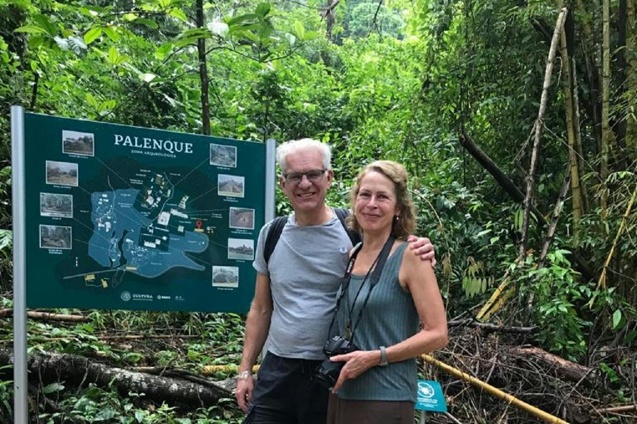 Two people standing in front of a map sign in a lush jungle environment, smiling at the camera.